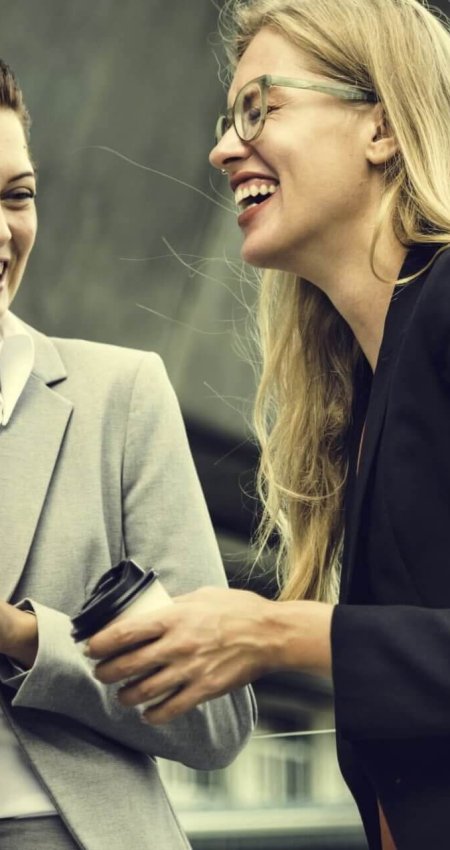 Două femei discutând și râzând, costume business.